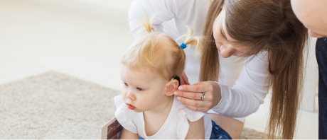 Woman bending down fixing the hearing aid in an infant girls left ear.