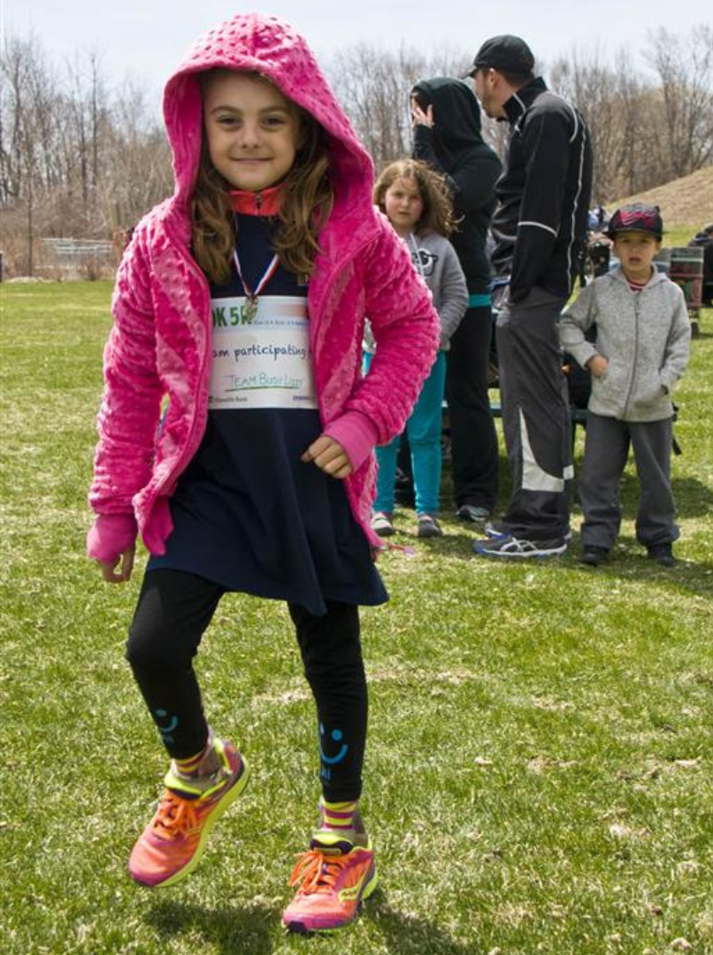 School-age Lizzy poses at the EOK 5K Run wearing a race bib that reads "Busy Lizzy"