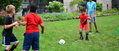 A boy in a red shirt is passing the ball to another boy in a black shirt, while a boy in a red shirt is trying to block the pass.