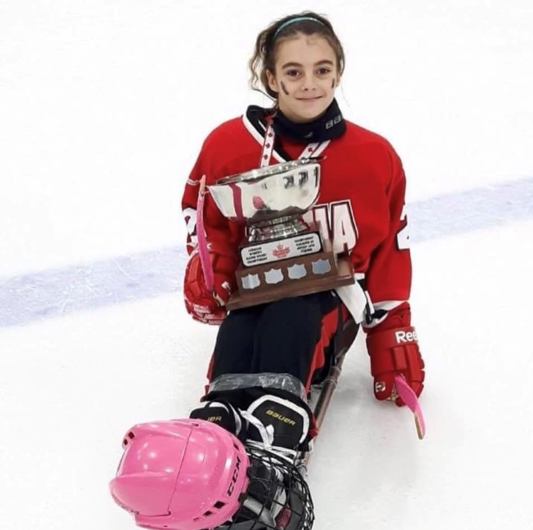 Lizzy dressed in ice skating attire on the rink, proudly holding the Women's Ontario Sledge Hockey provincial games trophy