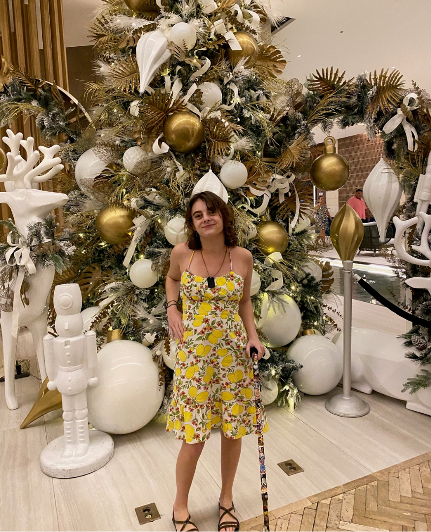 A recent photo of Lizzy: Lizzy poses in front of a large decorated Christmas tree wearing a yellow sundress and holding a cane.