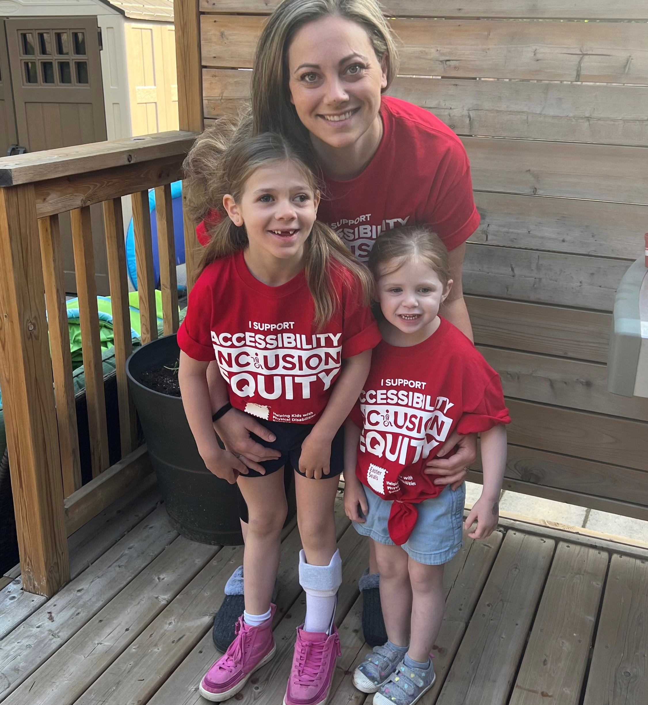 Mikayla poses with her mom, Amanda, and her little sister. They are wearing matching tshirts that read: "I support accessibility inclusion equity"