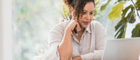 A woman watching a video on an open laptop leaning on her right hand and in her hand is a pencil