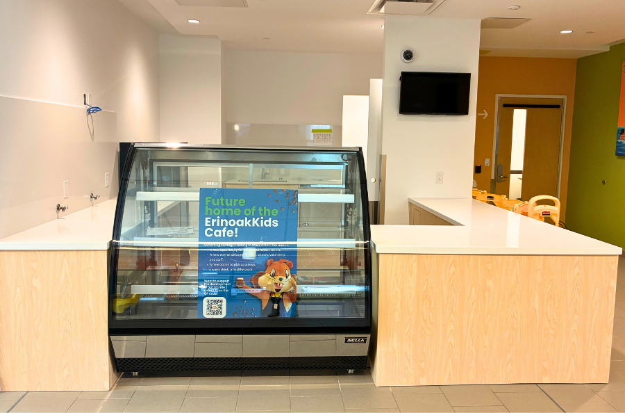 A close up of a cafe, with empty countertops and a display fridge with a sign inside that reads "future home of the ErinoakKids Cafe!"