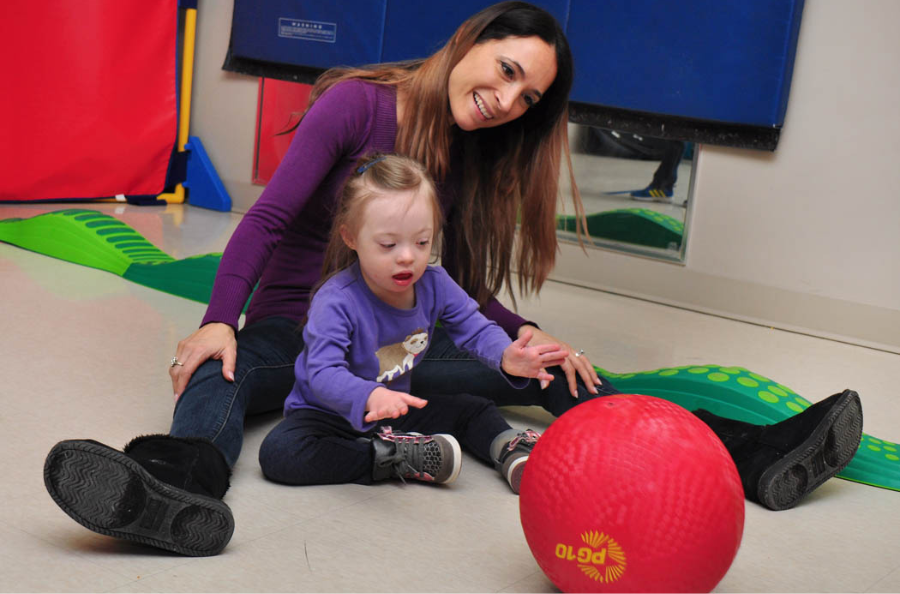 Adult sitting on the floor with a toddler rolling a red ball